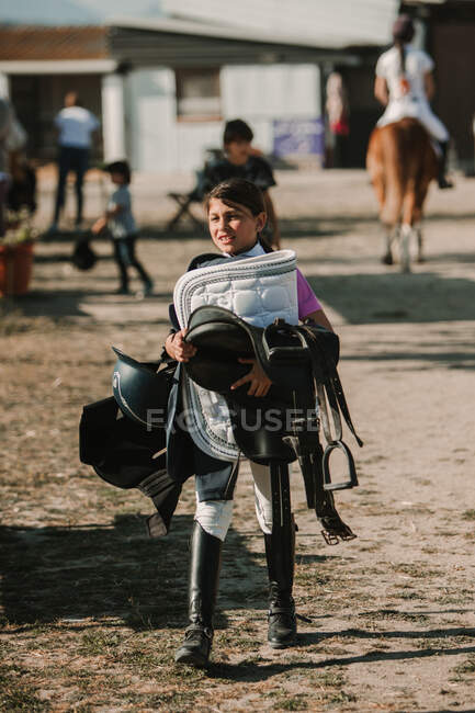 Little girl holding pile of jockey helmet with horse harness while walking on ranch in sunlight — Stock Photo