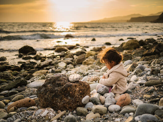 Cute interested toddler girl playing with stone while sitting on seaside — Stock Photo