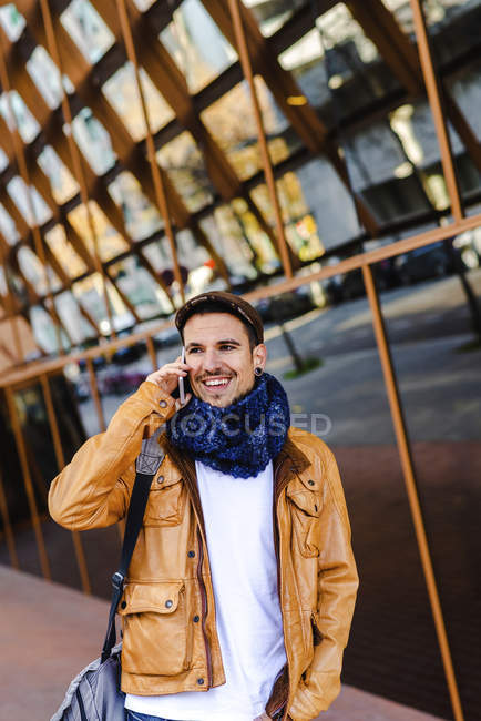 Модный мужчина в повседневной одежде с помощью мобильного телефона во время прогулки возле стеклянной стены современного здания — стоковое фото