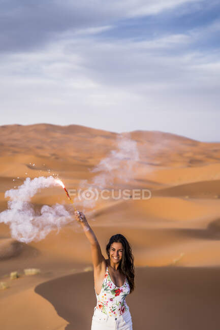 Cheerful stylish brunette holding arm raised with burning smoky firework standing in desert of Morocco — Stock Photo