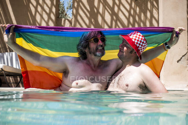 Мужчины среднего возраста с избыточным весом, прячущиеся от солнца под флагом ЛГБТ, сидя в бассейне на курорте — стоковое фото