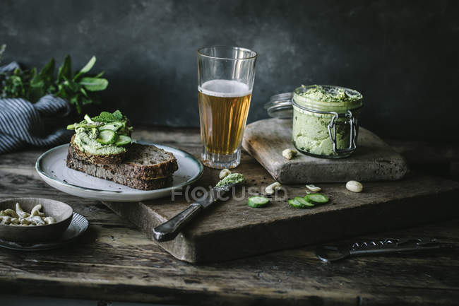 Тосты с зеленым кешью паштет, травы и ломтики огурца с банкой и стаканом пива на деревянной доске — стоковое фото