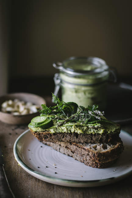 Toasts with green cashew pate, herbs and slices of cucumber with jar on wooden board — Stock Photo