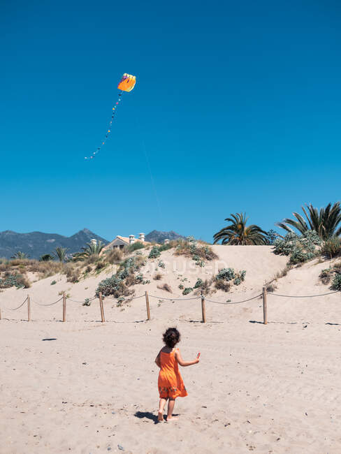 Visão traseira da criança pequena fêmea correndo à beira-mar com pipa voadora no fundo do céu azul — Fotografia de Stock