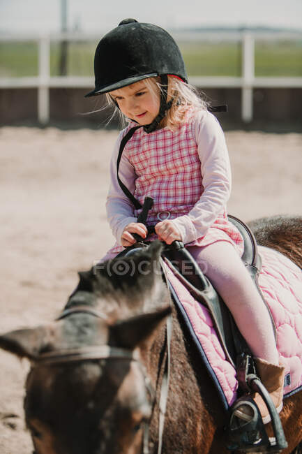 Ragazzina in abito e fieno di fantino seduta a cavallo mentre impara a cavalcare in pista — Foto stock