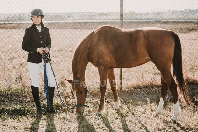 Adolescente en traje de jinete de pie con el caballo de castaño pastoreo cerca de la cerca de rancho - foto de stock