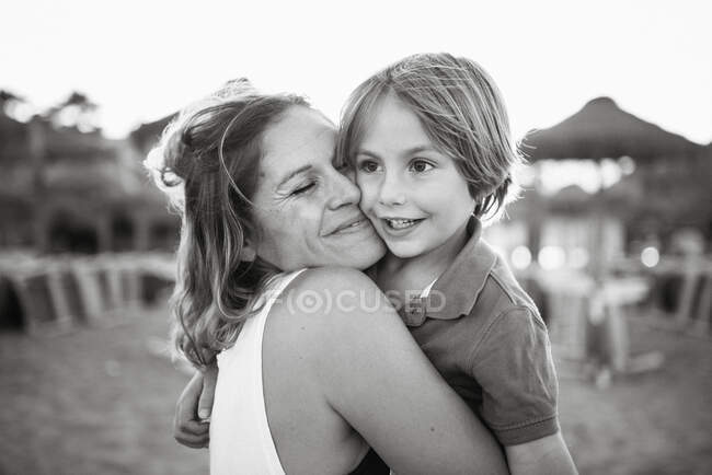 Mother embracing and kissing cute boy while standing together on beach in bright sunshine, black and white photo — Stock Photo