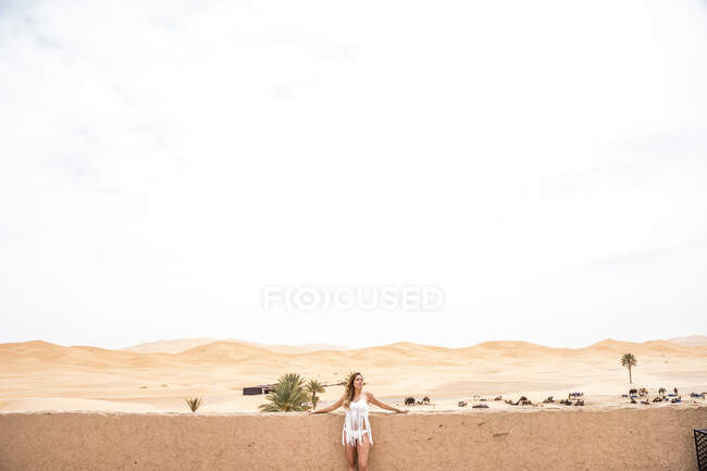 Красивая молодая женщина в белом топе опирается на стену, глядя в сторону бесконечной песчаной пустыни, Марокко — стоковое фото