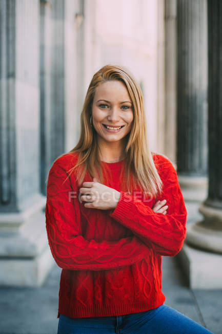 Cheerful young woman in trendy red sweater looking at camera while standing in front of marble columns on city street — Stock Photo