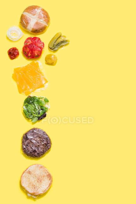 Different ingredients of a cheese burger wrapped in plastic on yellow background — Stock Photo