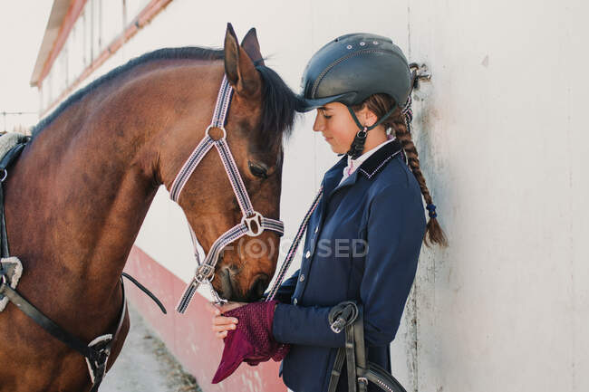 Side view of young teen woman in jockey helmet and jacket caressing horse standing together outdoors — Stock Photo