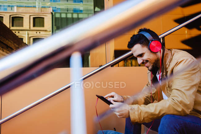 Hombre guapo en auriculares escuchando música y tableta de navegación mientras está sentado en las escaleras fuera del edificio moderno - foto de stock