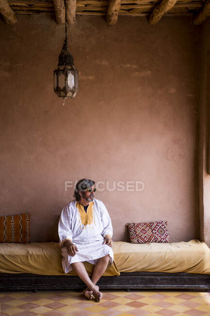 Hombre adulto en ropa larga sentado en el sofá en la terraza con valla de piedra en estilo oriental, Marruecos - foto de stock
