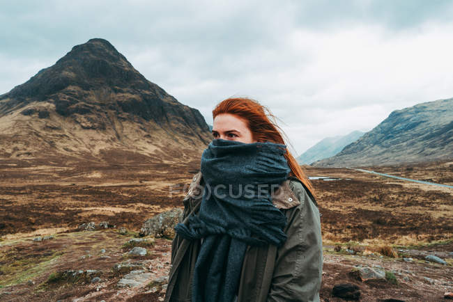 Hermosa mujer de jengibre envuelta en pañuelo de pie contra pintorescas montañas de Escocia - foto de stock