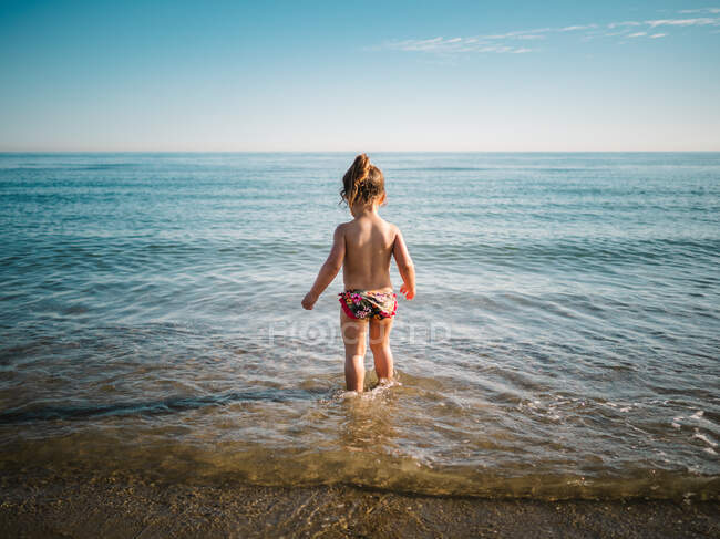 Back view of anonymous adorable toddler girl in swimming suit standing in warm water of calm sea — Stock Photo