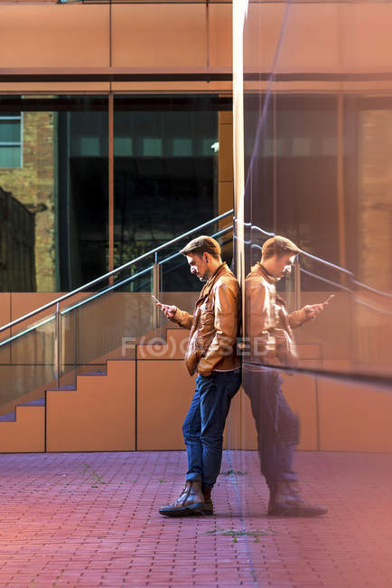Hombre en traje elegante usando el teléfono móvil mientras se apoya en la pared del edificio moderno en el día soleado - foto de stock