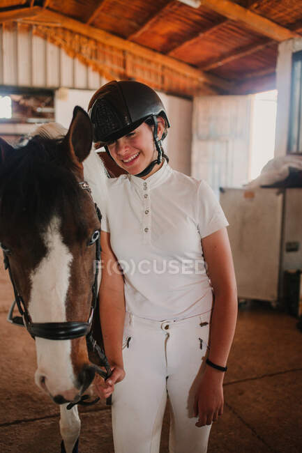 Jovem adolescente em roupa branca e capacete de jóquei levando cavalo fora de barraca para andar fora — Fotografia de Stock