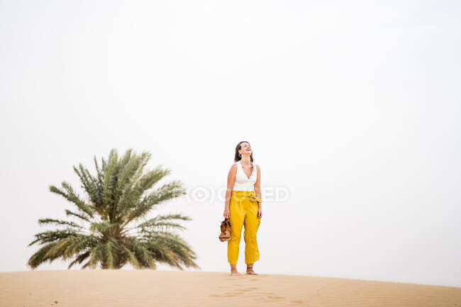 Alegre mujer rubia elegante sosteniendo zapatos mientras camina en el desierto de Marruecos - foto de stock