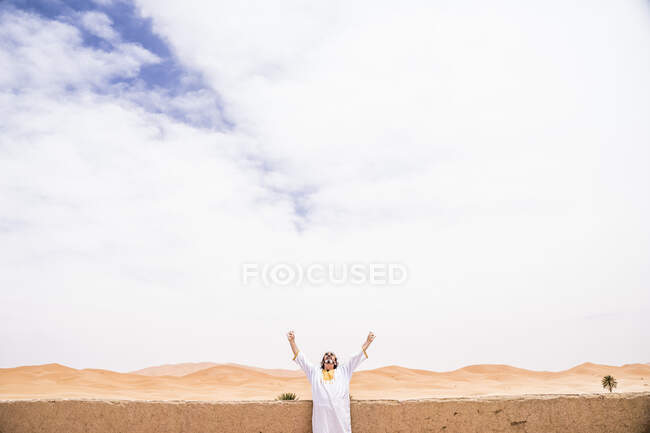 Евфорический взрослый мужчина смотрит на небо с распростертыми руками возле каменного забора на террасе против бесконечной пустыни, Марокко — стоковое фото