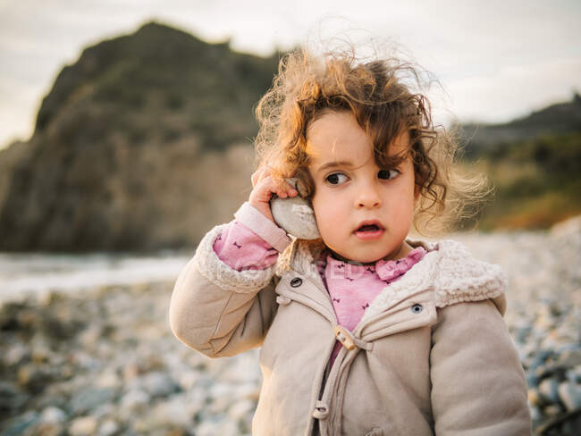 Retrato de linda niña escuchando concha con atención arrebatada mientras descansa en la playa pedregosa - foto de stock