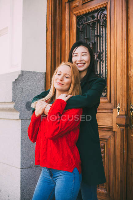 Glückliche asiatische Frau lächelnd und wegschauend, während sie vor der Ziertür eines gealterten Gebäudes steht und ihren kaukasischen Freund umarmt — Stockfoto