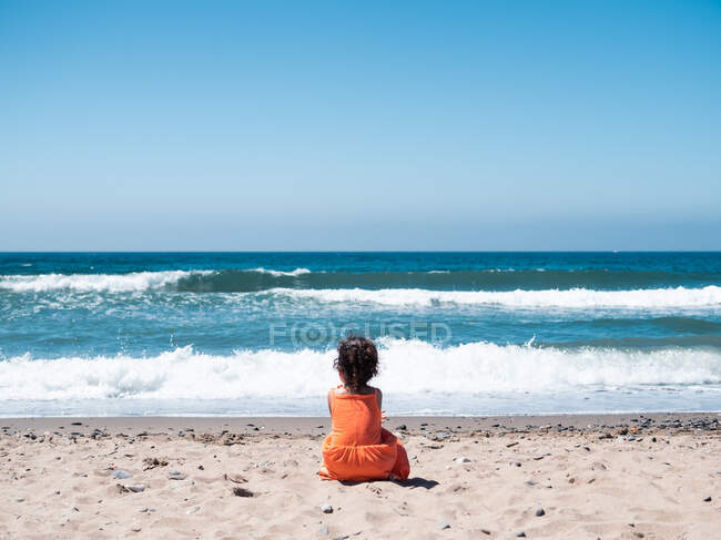 Vue arrière de l'enfant femelle en robe orange assis sur le bord de mer sablonneux et regardant la mer et le ciel — Photo de stock