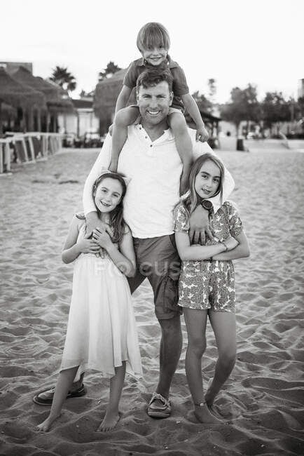 Adult man with laughing boy on shoulders standing with beautiful little girls on beach looking at camera, black and white photo — Stock Photo