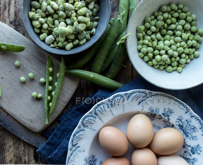 Сверху веревки свежий зеленый горох в миске и яйца на деревянном столе во время приготовления пищи — стоковое фото