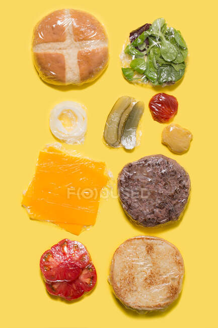Diferentes ingredientes de um hambúrguer de queijo envolto em plástico sobre fundo amarelo — Fotografia de Stock