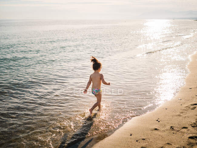Back view of anonymous cute female kid in swimming slips running on sandy beach against calm sea — Stock Photo