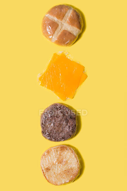 Різні інгредієнти сирного бургеру, загорнуті в пластик на жовтому тлі — стокове фото