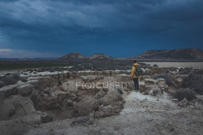 Vista laterale del giovane in giacca gialla e zaino in piedi sulla pietra e guardando impressionante cielo blu al crepuscolo in semi-deserto Bardenas Reales Navarra Spagna — Foto stock