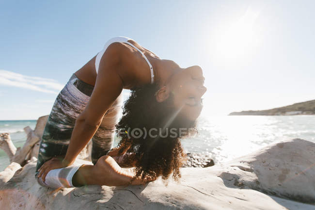 Primer plano de mujer joven atractiva afroamericana que está parada en el puente de la postura del yoga en el fondo del agua calma en día soleado - foto de stock