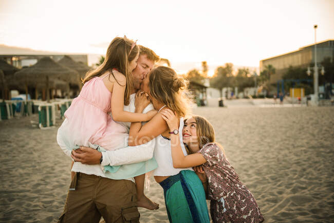 Adulto amante hombre y mujer besándose mientras sostiene un abrazo con hijas de pie juntos en la playa en la espalda iluminada - foto de stock