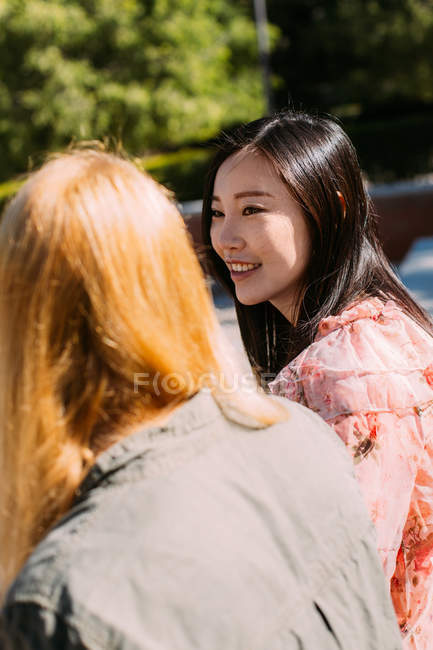 Young women in casual outfits talking and looking at each other while sitting on bench in park — Stock Photo