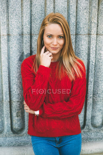 Young woman in trendy red sweater touching neck and looking at camera while leaning on marble wall on city street — Stock Photo
