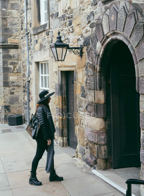 Vista lateral de la mujer de moda en traje negro y sombrero caminando por la calle antigua pavimentada con edificio de piedra, Escocia - foto de stock