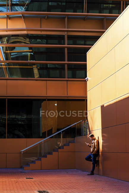 Homem em roupa elegante usando telefone celular enquanto se inclina na parede do edifício moderno no dia ensolarado — Fotografia de Stock