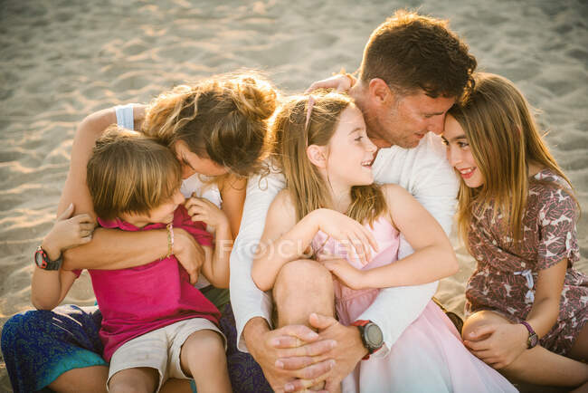 Adulto amorevole uomo e donna con figlio allegro e figlie seduti insieme guardando l'un l'altro sulla spiaggia in retroilluminazione — Foto stock