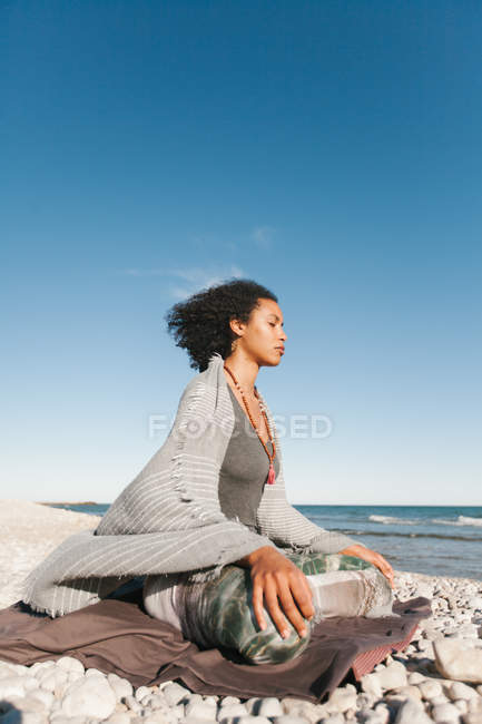 Афроамериканская молодая женщина медитирует в позе йоги лотоса на песчаном пляже в светлый день — стоковое фото
