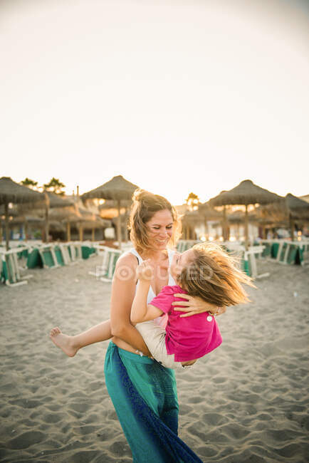 Seitenansicht einer lachenden Frau, die ihren fröhlichen, verspielten Sohn auf Händen trägt, während sie im Sonnenuntergang am Strand steht — Stockfoto