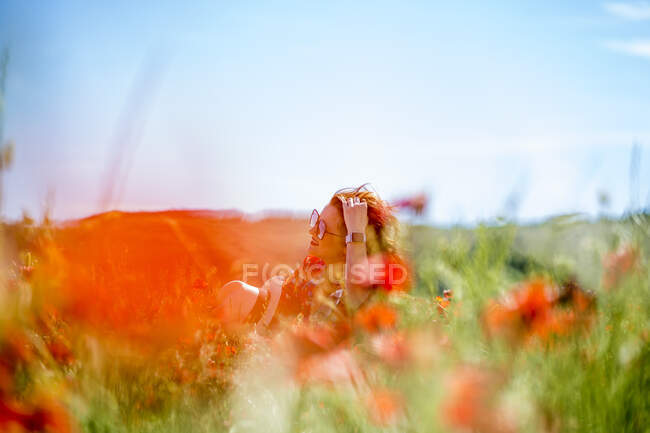 Stylish young female touching hair and looking away while sitting on ground in blooming meadow on sunny summer day — Stock Photo
