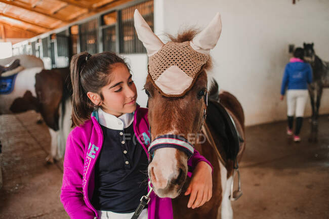 Маленькая девочка обнимает маленького пони в милой шляпе на ушах, стоящих внутри конюшни — стоковое фото