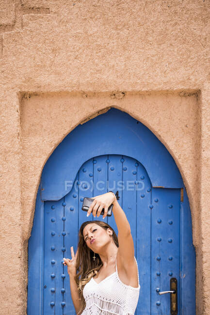 Joven alegre vistiendo top blanco con bikini tomando selfie con teléfono contra puerta oriental azul en pared de piedra, Marruecos - foto de stock