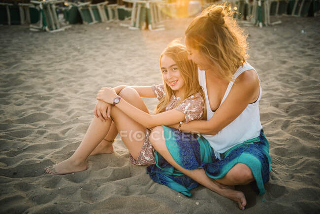 Mujer adulta abrazando hermosa chica con amor en la playa de arena en la luz del atardecer - foto de stock