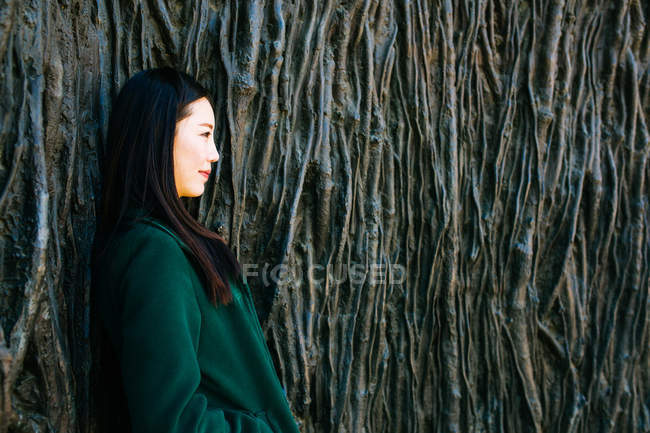 Nachdenkliche Asiatin im trendigen Outfit schaut weg, während sie sich an die Wand lehnt, mit Relief von Baumwurzeln — Stockfoto