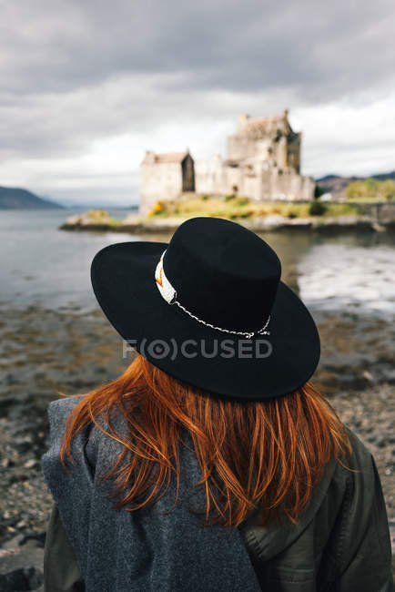 Rückseite einer stilvollen Frau mit Hut, die eine alte Steinburg an der Küste in den Bergen in Schottland betrachtet — Stockfoto