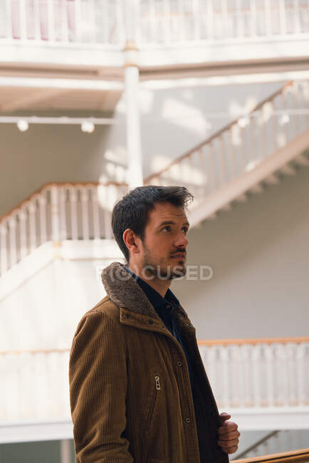 Взрослый мужчина в коричневой куртке с руками в карманах, стоящий в светлом зале с лестницей, смотрящий в камеру — стоковое фото
