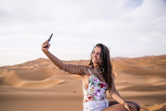 Молодая женщина делает селфи с телефоном посреди песчаной пустыни, Марокко — стоковое фото