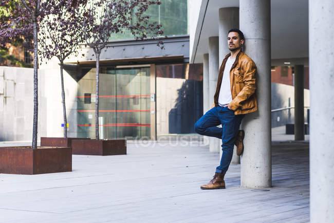 Confident trendy guy leaning on concrete pillar near leather couch outside contemporary building on city street — Stock Photo